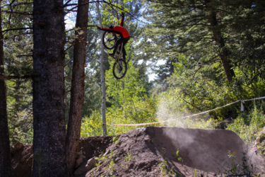 A mountain biker performs a jump over a dirt ramp, soaring through the air with a forest backdrop. Dust is seen rising from the ramp, and trees surround the area, creating a natural setting for biking activities. Deer Jumps mountain bike trail.