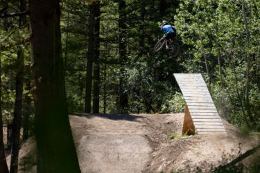 A mountain biker performing a jump off a wooden ramp in a forested area, surrounded by trees and sunlight. The biker is mid-air, showcasing skills in a natural outdoor setting. Deer Jumps mountain bike trail.