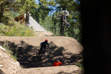 A mountain biker performing a jump on a dirt trail with two other riders in the foreground, surrounded by trees and natural scenery. Deer Jumps mountain bike trail.