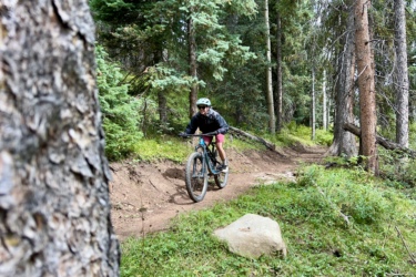 A mountain biker riding along a dirt trail surrounded by trees and greenery. The biker is wearing a helmet and a jacket, navigating a curved path with a large rock visible on the side. Cutthroat Trail mountain bike trail.
