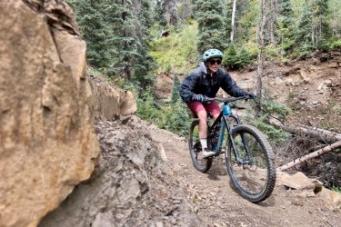 A person riding a mountain bike on a narrow trail surrounded by green trees and rocky terrain. The rider is wearing a helmet, sunglasses, and a jacket, navigating the path with focus. Cutthroat Trail mountain bike trail.