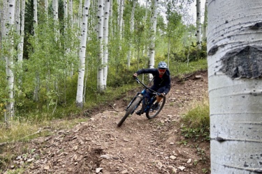 A mountain biker leans into a turn on a dirt trail surrounded by tall aspen trees. The cyclist is wearing a helmet and a dark outfit, navigating the rugged terrain with a focused expression. The lush greenery and white bark of the aspens create a vibrant backdrop. Cutthroat Trail mountain bike trail.