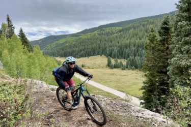A cyclist in a blue helmet and dark jacket rides a mountain bike along a rocky path with a lush green landscape in the background, featuring tall trees and a winding road below. The sky is partly cloudy, suggesting an outdoor adventure in a mountainous area. Cutthroat Trail mountain bike trail.