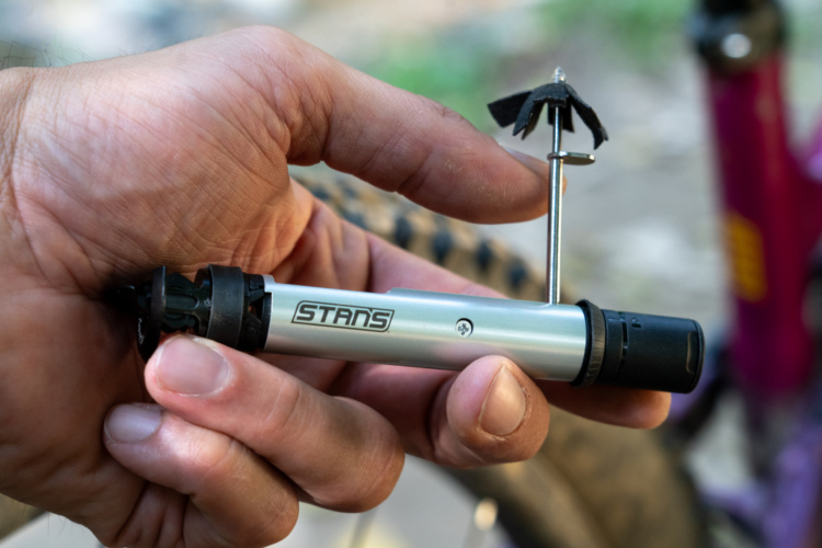 A close-up image of a hand holding a silver Stans Incredible DART tool with a black nozzle and a small tool attached at the end. The inflator features the brand name "STANS." The background is slightly blurred, focusing on the inflator and hand.