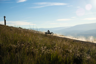 A dirt bike rider is ascending a grassy hillside against a backdrop of blue sky and distant mountains, creating a trail of dust in the air. Deepest Darkest mountain bike trail.