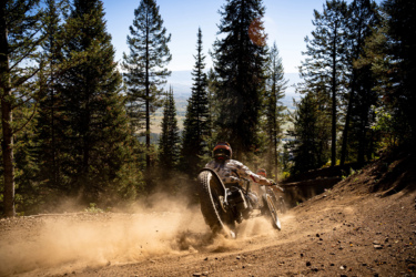 A person riding a three-wheeled off-road vehicle on a dirt trail surrounded by tall evergreen trees. Dust is being kicked up from the ground as the vehicle speeds through the forest, with a scenic view of a valley in the background. The sky is clear and blue. Deepest Darkest mountain bike trail.