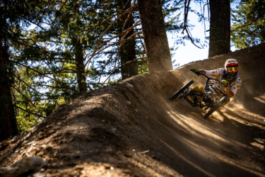 A person riding a mountain bike on a dirt trail in a wooded area, leaning into a turn as dust is kicked up from the ground. Tall trees and sunlight filtering through the leaves create a dynamic outdoor scene. Deepest Darkest mountain bike trail.