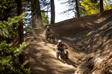 Two riders in specialized off-road carts navigate a dusty trail surrounded by trees. The first rider, wearing an orange helmet and plaid shirt, races ahead on a downhill path, while the second rider in a white helmet and patterned shirt follows closely behind. Sunlight filters through the trees, creating a dynamic outdoor scene filled with dust and movement. Deepest Darkest mountain bike trail.