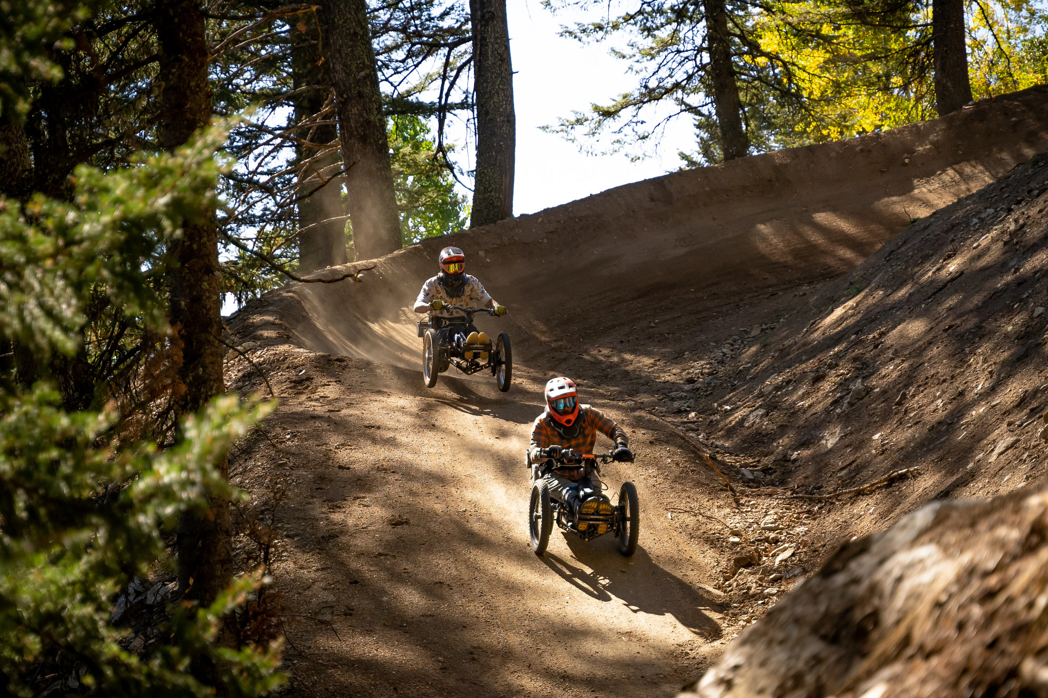 Two riders in specialized off-road carts navigate a dusty trail surrounded by trees. The first rider, wearing an orange helmet and plaid shirt, races ahead on a downhill path, while the second rider in a white helmet and patterned shirt follows closely behind. Sunlight filters through the trees, creating a dynamic outdoor scene filled with dust and movement. Deepest Darkest mountain bike trail.