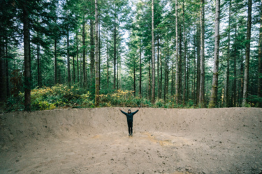 A person stands with arms raised in a large, circular dirt area surrounded by tall trees and dense forest greenery. The scene depicts a tranquil outdoor setting with autumn foliage visible in the background. Blue Ribbon mountain bike trail.