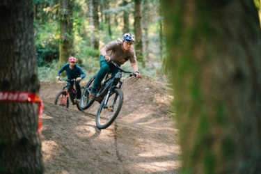 Two mountain bikers riding through a forest trail. One rider, in a brown shirt and helmet, is jumping over a dirt mound, while the other, wearing a blue shirt and red helmet, follows closely behind. The scene is surrounded by lush green trees, highlighting the natural outdoor setting. Blue Ribbon mountain bike trail.
