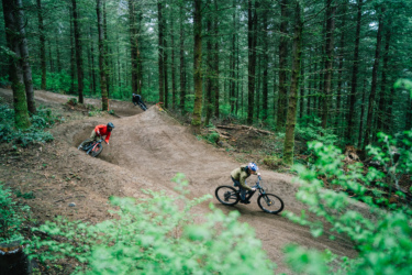 Three mountain bikers navigate a winding dirt trail through a lush forest, surrounded by tall trees and greenery. The riders are leaning into the curves, showcasing their skills as they enjoy the outdoor setting.
