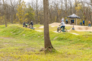 A group of children and adults riding bikes on a dirt track, with grassy mounds and a wooded background. A tent is set up nearby with people gathered around, enjoying the outdoor activity. Palmer Urban Mountain-bike Park (The PUMP) mountain bike trail.