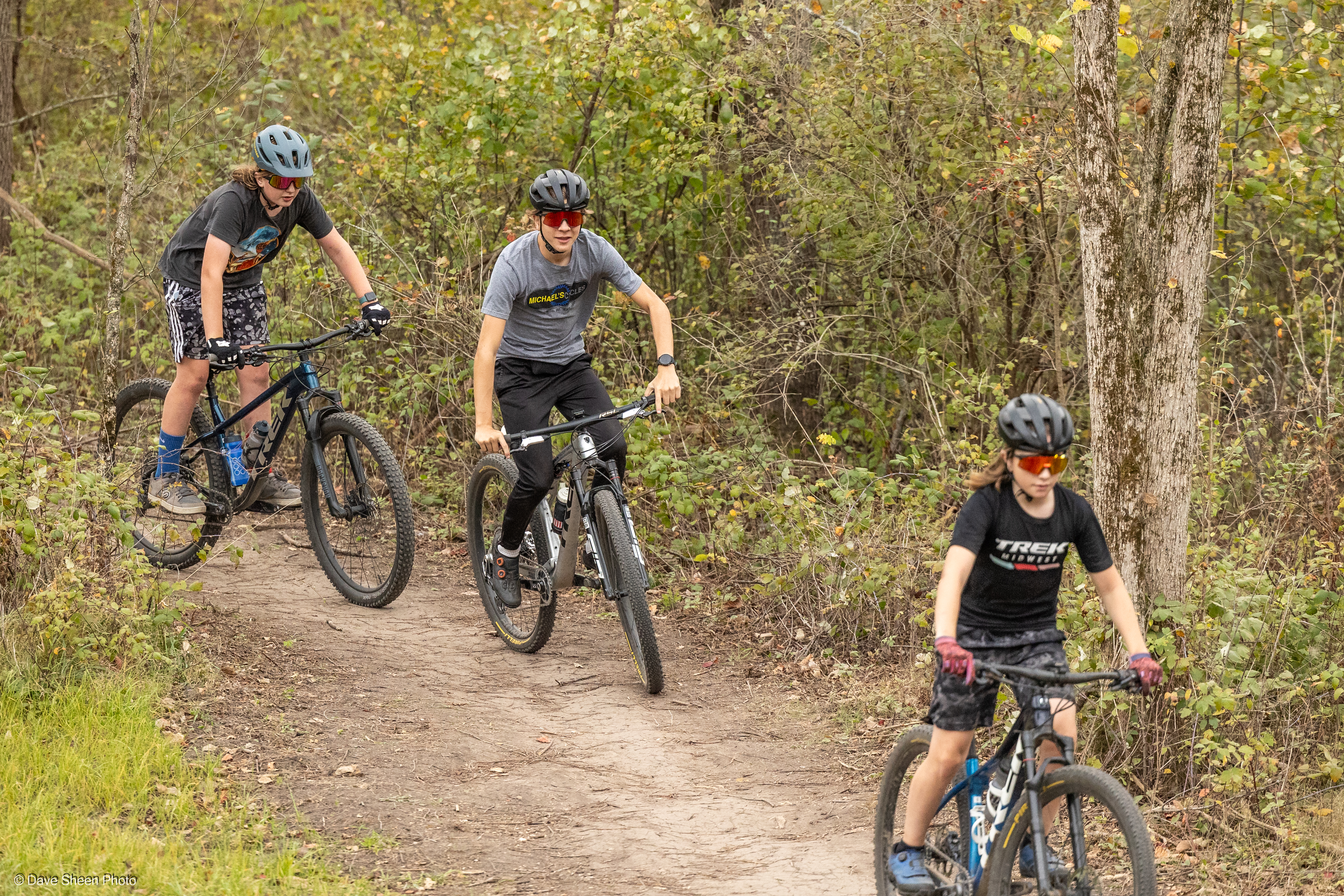 Three young cyclists ride their mountain bikes along a dirt trail in a wooded area. They are wearing helmets and sunglasses, and the surroundings are filled with greenery and trees. The scene captures a dynamic moment of outdoor recreation and adventure. Palmer Urban Mountain-bike Park (The PUMP) mountain bike trail.