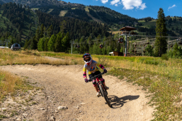 A child riding a mountain bike on a dirt trail in a scenic outdoor area, surrounded by lush greenery and mountains. The child is wearing a helmet and protective gear, and the background features a ski lift with gondolas in operation. The sky is blue with a few clouds, indicating a bright, sunny day. Lucky Charm mountain bike trail.