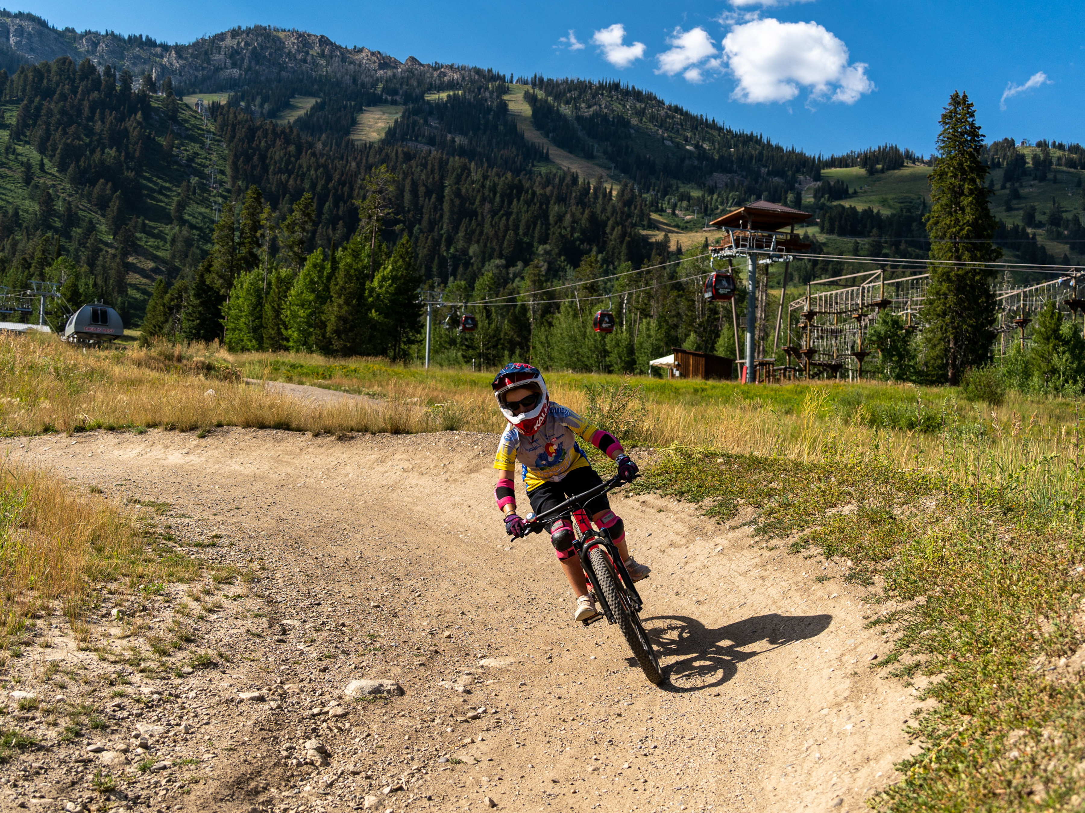 A child riding a mountain bike on a dirt trail in a scenic outdoor area, surrounded by lush greenery and mountains. The child is wearing a helmet and protective gear, and the background features a ski lift with gondolas in operation. The sky is blue with a few clouds, indicating a bright, sunny day. Lucky Charm mountain bike trail.