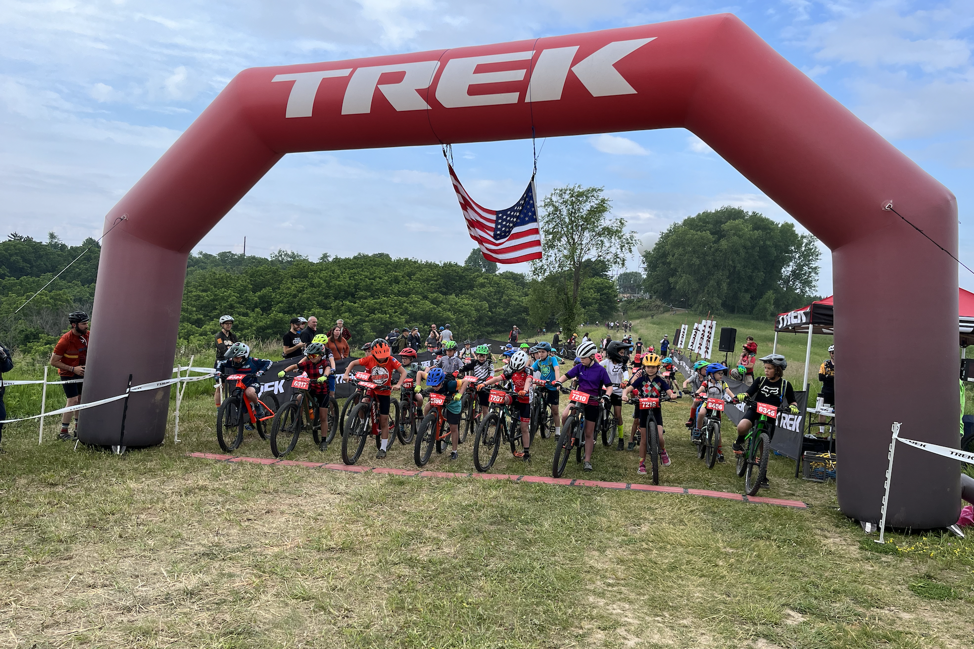 A group of young cyclists lined up at the starting line of a mountain biking race, beneath a large red inflatable arch displaying the Trek logo and an American flag hanging above. The scene is set in a grassy area with trees in the background, and there are spectators and event banners visible in the distance.