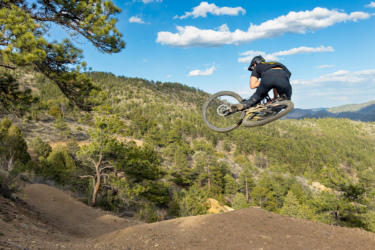 A mountain biker performs a jump off a dirt ramp, showcasing an impressive airborne maneuver against a backdrop of lush green hills and a clear blue sky with scattered clouds. Drop Shaft mountain bike trail.