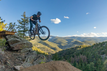A mountain biker in mid-air jumps off a rocky ledge with a mountainous landscape in the background, showcasing trees and valleys under a clear blue sky with a few clouds.