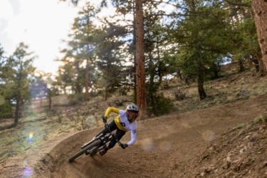 A mountain biker in a yellow long-sleeve shirt leans into a dirt turn on a forest trail, surrounded by trees and a clear sky. Dust is kicked up as the rider navigates the curved path, showcasing a dynamic action shot in a natural setting.