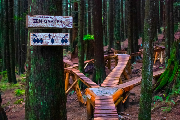 Wooden signpost reading "Zen Garden" with a decorative lotus symbol, situated among tall, dark trees in a forest. A winding wooden pathway is visible in the background, leading into the tranquil, natural environment.