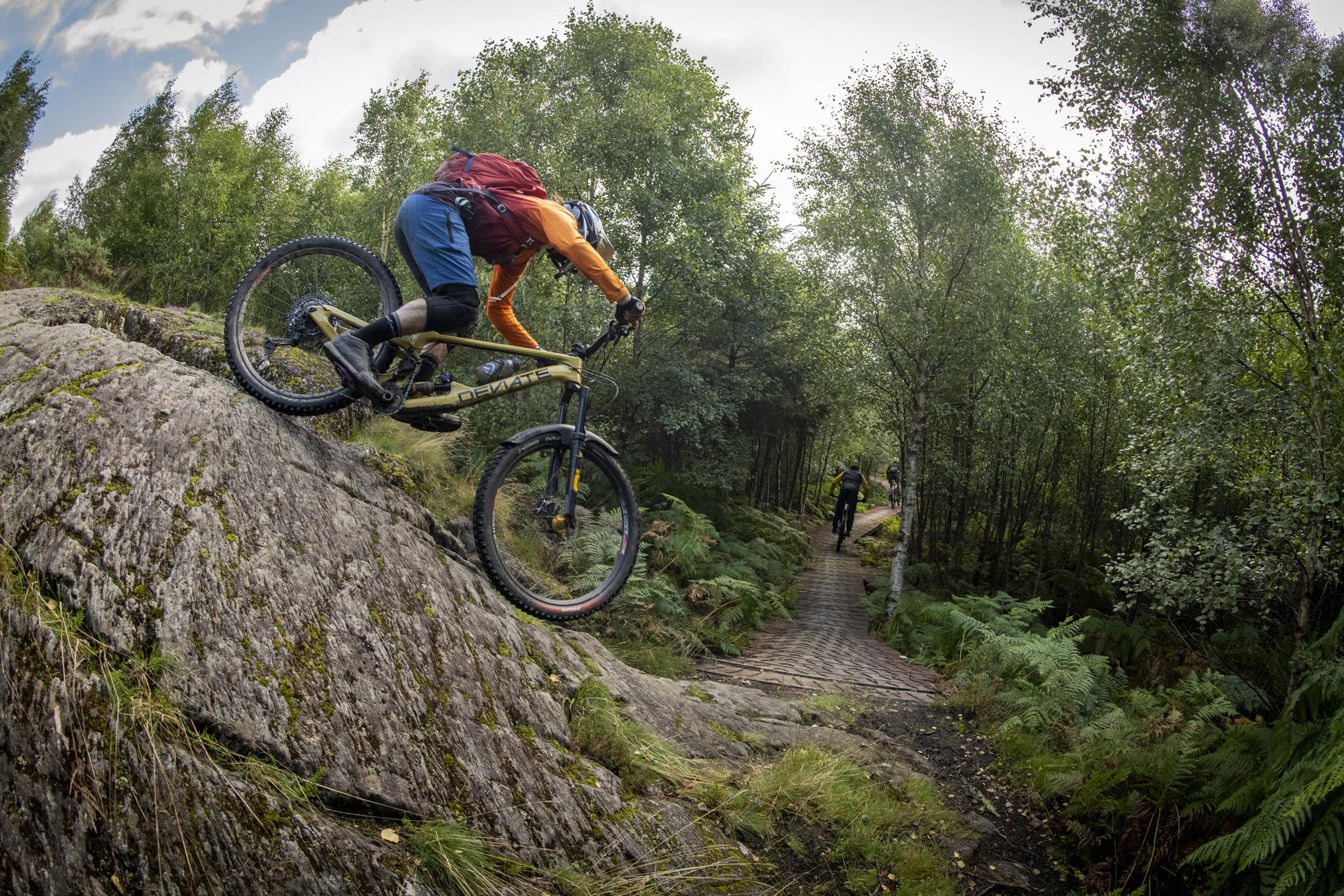 A mountain biker navigating a rocky terrain with lush greenery and trees surrounding a winding path in the background. The biker wears a helmet and bright clothing, showcasing an adventurous moment in nature. Comrie Croft Trails mountain bike trail.
