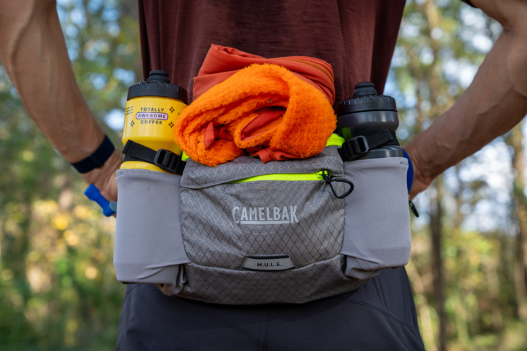 A close-up view of a gray CamelBak M.U.L.E. pack worn around the waist, featuring a vibrant orange fleece, a yellow water bottle labeled "TOTALLY AWESOME COFFEE," and a dark-colored water bottle on either side. The background shows a blurred forest setting, enhancing the outdoor theme.