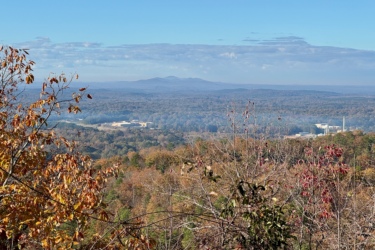 Scenic view of rolling hills and distant mountains under a clear blue sky, with autumn foliage in the foreground showcasing shades of orange, yellow, and red. A valley below features a few structures surrounded by trees. Bomb Dog mountain bike trail.