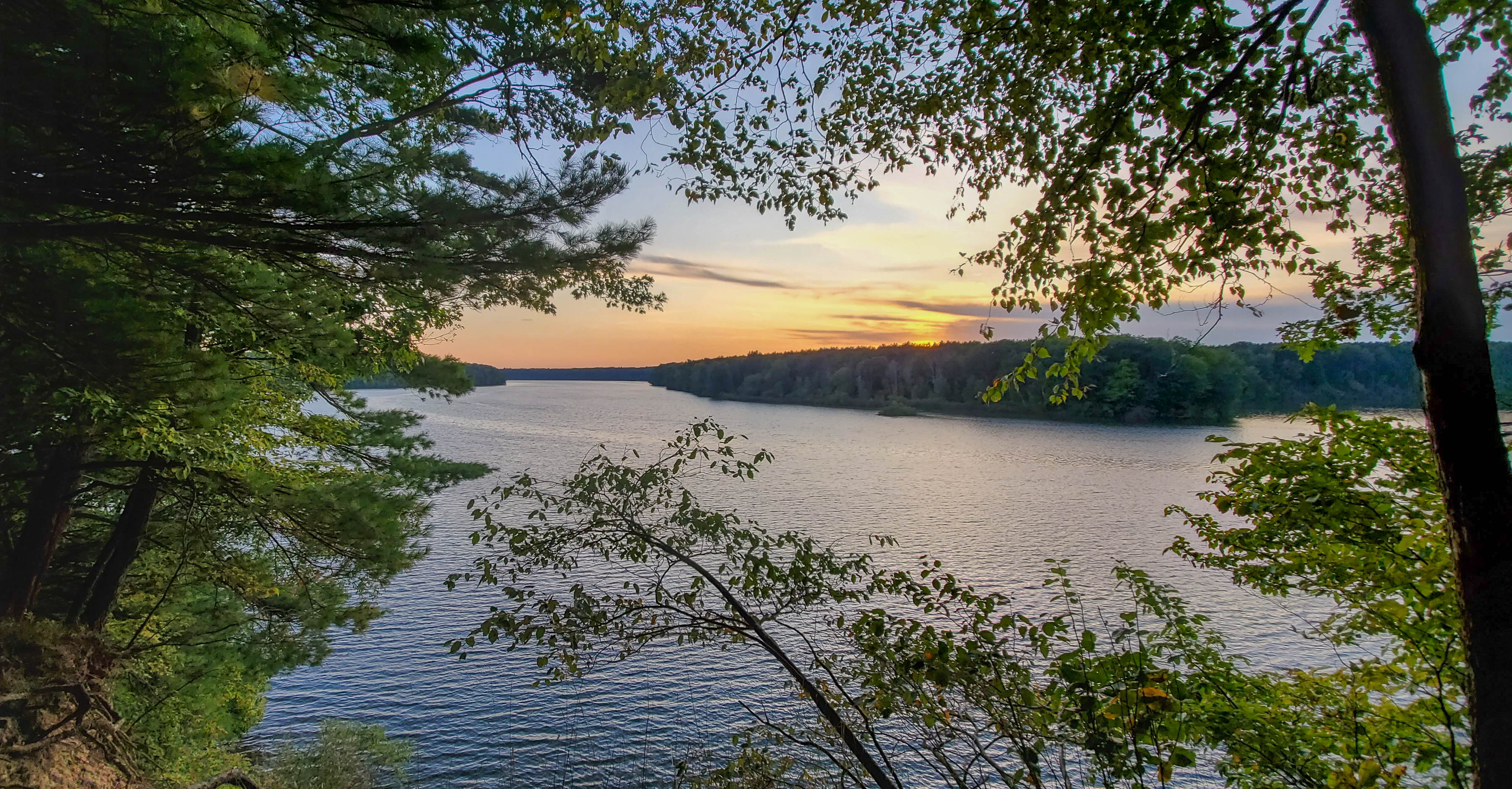 A serene view of a river surrounded by lush greenery, with the sun setting behind distant trees. The calm water reflects the warm colors of the sunset, creating a peaceful natural scene framed by branches and leaves. The Dragon mountain bike trail.