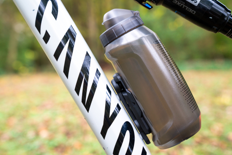 A close-up of a bicycle frame with the word "CANYON" prominently displayed, featuring a brown water bottle securely mounted on the frame. The background is blurred, showcasing a natural setting with greenery and soft foliage.