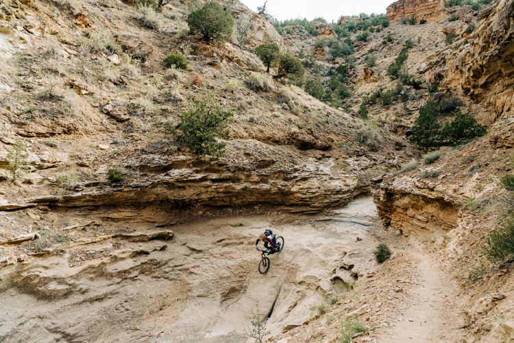 A mountain biker performing a jump in a rocky canyon surrounded by dry hills and sparse vegetation. The scene captures the dynamic movement of the rider mid-air, highlighting rugged terrain and natural geological features.