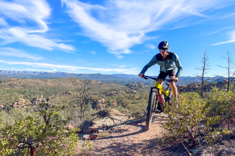 A person riding a mountain bike on a trail surrounded by rocky terrain and greenery, with a scenic view of hills and a blue sky with wispy clouds in the background.