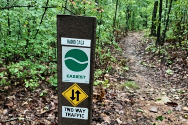 A trail sign in a wooded area, indicating the "Radio Gaga" trail, marked as the easiest level. The sign includes symbols for two-way traffic and directions for hikers, with a view of a dirt path leading into the greenery. The ground is covered with fallen leaves and the surroundings are lush with trees. Radio Gaga mountain bike trail.
