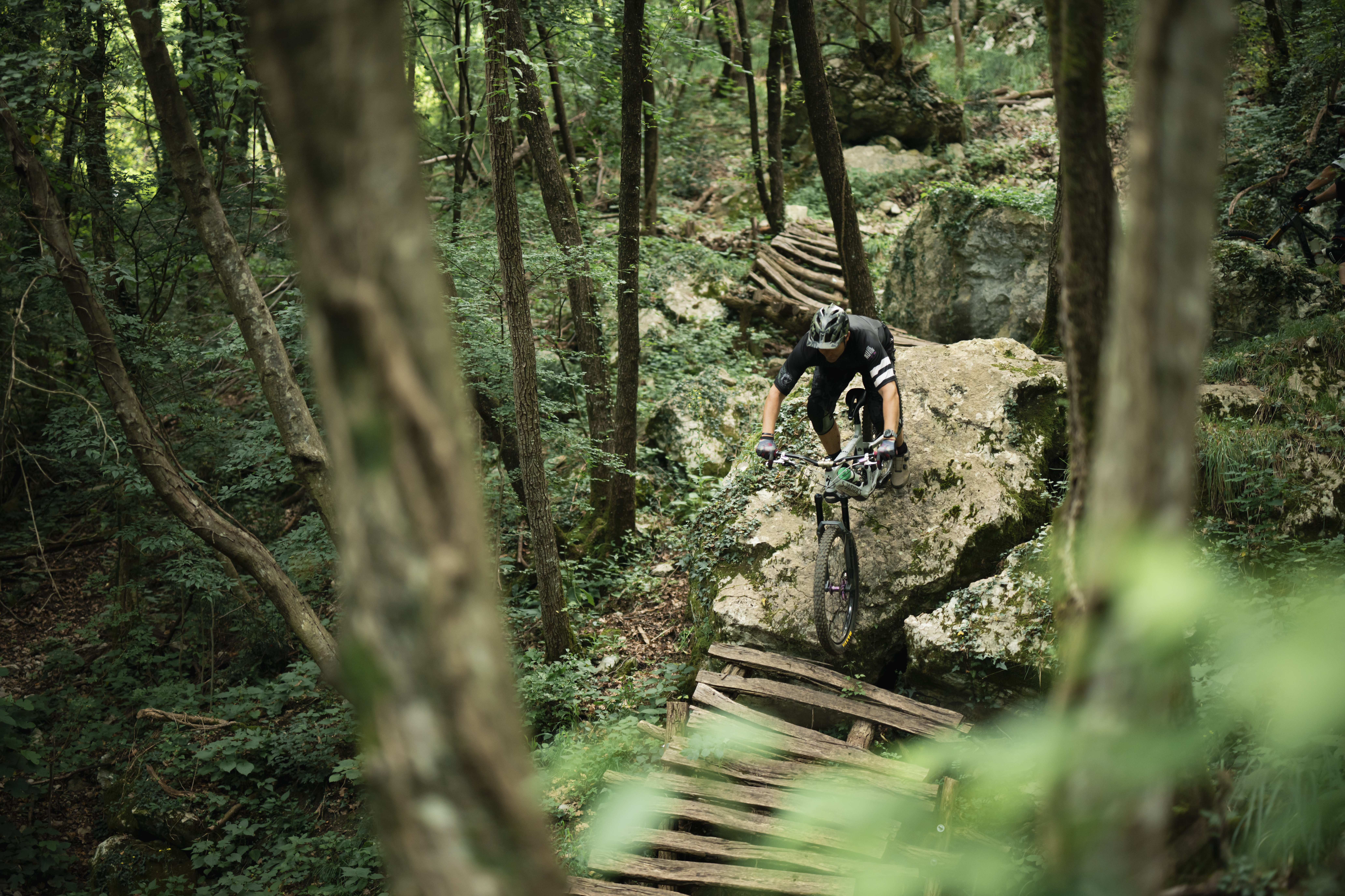 A mountain biker navigating a rocky terrain on a forest trail, balancing on a wooden bridge surrounded by green foliage and trees. The cyclist is focused and wearing protective gear, showcasing the thrill of outdoor biking. Carovnica - The Witch // TCS mountain bike trail.