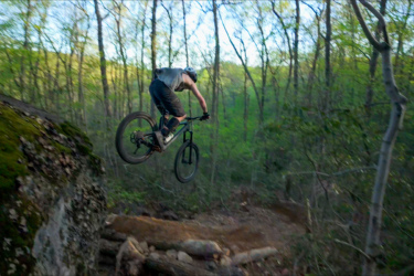 A mountain biker performing a jump off a rocky outcrop in a lush forest, with trees and green foliage in the background. The bike's wheels are elevated off the ground as the rider leans forward, showcasing action and adventure in nature.