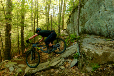 A mountain biker maneuvering over rocky terrain in a forested area, with trees and leaves visible in the background. The rider is crouched low on the bike, wearing a helmet and black clothing, showcasing an action shot as they navigate a challenging section of the trail. Satan's Ridge mountain bike trail.
