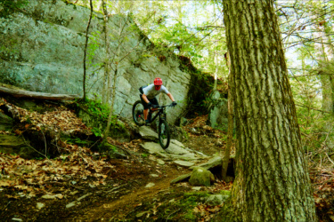 A person wearing a red helmet is riding a mountain bike down a rocky trail in a forested area. Surrounding the trail are trees and patches of leaves on the ground, with a large rock face in the background. Satan's Ridge mountain bike trail.