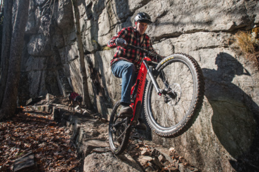 A mountain biker in a black and red plaid shirt and helmet skillfully navigates a rocky trail, lifting the front wheel of a red bike off the ground. In the background, a dog in a matching plaid outfit explores the trail amidst fallen leaves and rocky terrain, with a stone cliff rising on one side. Satan's Ridge mountain bike trail.