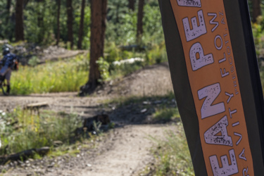 A close-up of a banner displaying "Bean Peaks Gravity Flow Trails" in a wooded area, with a dirt path visible in the background. The banner is prominently featured on the left side, and trees and greenery surround the trail. Bean Peaks mountain bike trail.