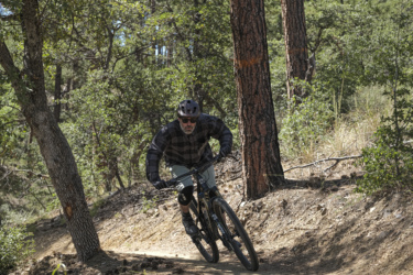 A person mountain biking on a dirt trail surrounded by trees, wearing a helmet and sunglasses, dressed in a plaid shirt and shorts, focused on navigating the terrain. Bean Peaks mountain bike trail.