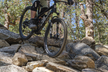 A cyclist navigates rocky terrain on a mountain bike, showcasing skill and balance. The scene is set in a forest with tall trees and clear blue skies, highlighting the adventurous spirit of outdoor biking. Bean Peaks mountain bike trail.