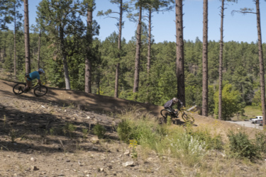 Two mountain bikers navigate a dirt trail through a forest of tall pine trees. The first rider, dressed in a blue shirt and a helmet, leans into a curve on the left, while the second rider, wearing a dark jacket and a helmet, follows closely behind. The environment is sunny with greenery and wildflowers along the trail. In the background, a glimpse of a vehicle is visible parked near the tree line. Bean Peaks mountain bike trail.