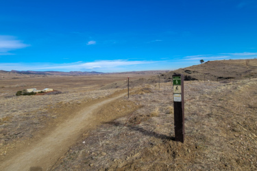 A dirt path leading into open terrain with sparse vegetation, flanked by fences and a trail marker indicating walking and biking routes. The background features rolling hills under a clear blue sky. A small building can be seen at a distance among the grassland. Boulder Valley Ranch mountain bike trail.
