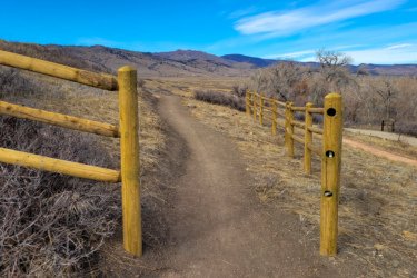 A dirt path bordered by wooden fencing, winding through a grassy landscape with dry vegetation and mountains in the background under a clear blue sky. The scene suggests a serene natural setting ideal for walking or hiking. Boulder Valley Ranch mountain bike trail.