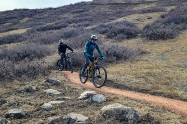 Two cyclists riding mountain bikes along a dirt path in a hilly, grassy area. The landscape features sparse vegetation and rocky outcroppings, with a cloudy sky overhead. One cyclist is in the foreground, wearing a blue jacket and gloves, while the other is further back, dressed in a dark jacket. Boulder Valley Ranch mountain bike trail.
