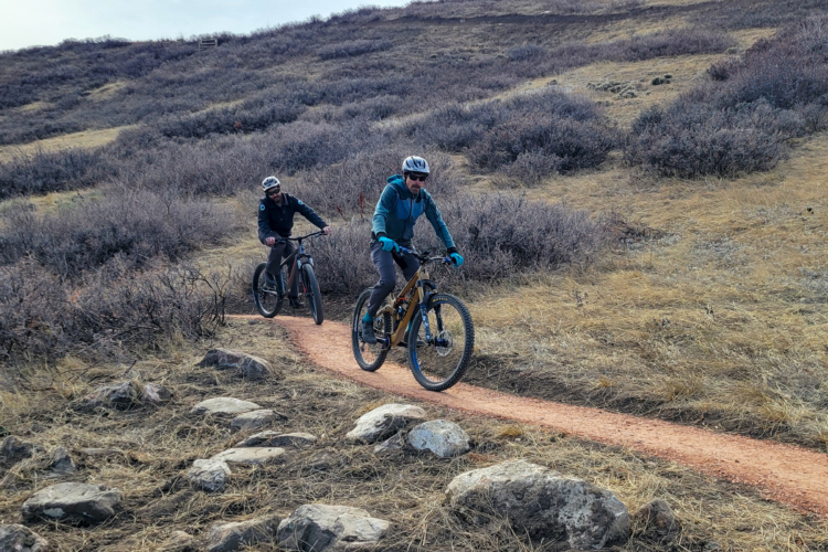 Two mountain bikers riding along a dirt trail surrounded by dry grass and shrubs. One cyclist is in the foreground wearing a blue jacket and helmet, while the other is further back in a black jacket. The trail features some rocks along the sides and slopes gently upward in the background.