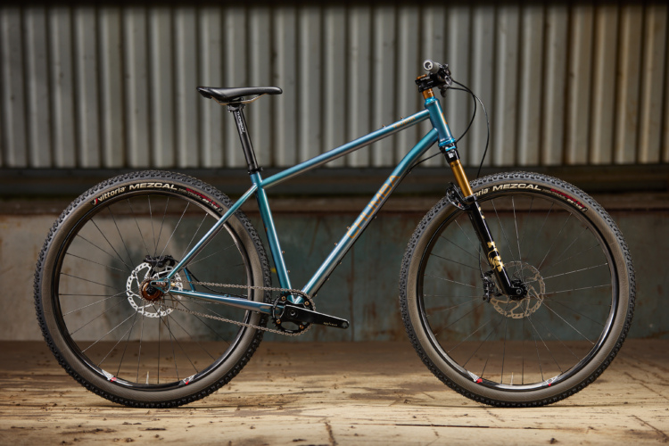 A sleek, blue mountain bike is positioned on a wooden floor against a backdrop of corrugated metal. The bike features a sturdy frame, thick tires with a tread pattern suited for off-road riding, and high-quality components including disc brakes. The overall design showcases a blend of durability and style, ideal for outdoor adventures.