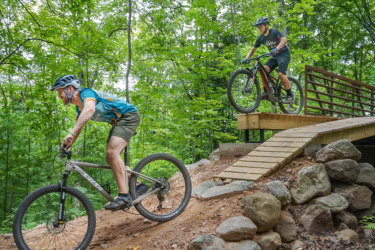 Two mountain bikers navigating a dirt trail in a lush, green forest. One rider is descending a slope, while the other is jumping off a small wooden bridge. Both are wearing helmets and athletic gear, immersed in their biking activity. The scene showcases the excitement of outdoor biking in a natural setting. WinMan Trails mountain bike trail.