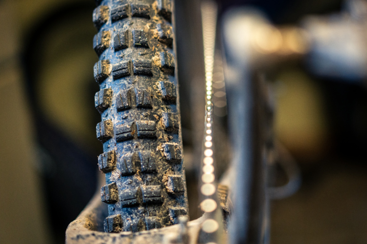 Close-up shot of a WTB Judge SG1 bicycle tire with a rugged tread pattern, showing dirt and debris, alongside a blurred bicycle chain in the background.