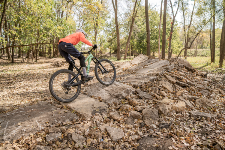 A mountain biker in an orange jacket and helmet skillfully navigates a rocky terrain covered in leaves, surrounded by trees in a natural setting.